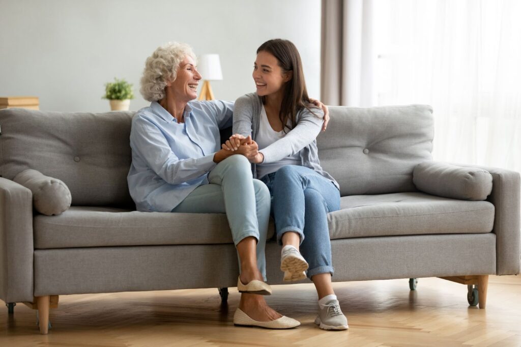 A senior woman and her daughter smiling while holding hands on a couch while talking about senior living.