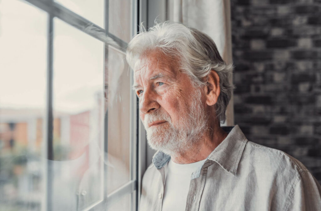 An older adult stands at a window and looks outside pensively.