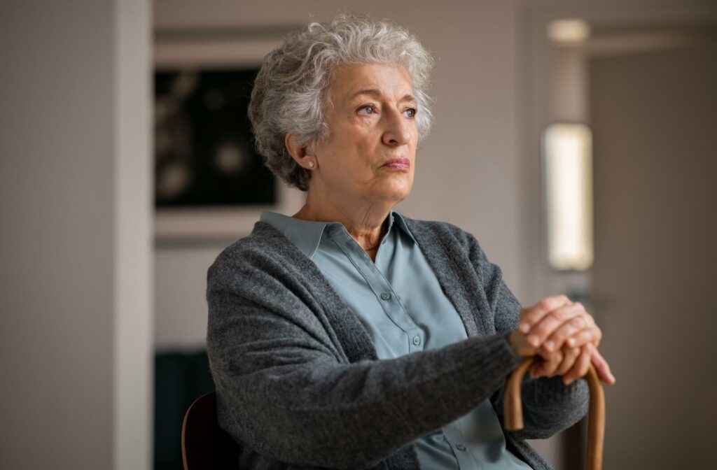An irritated older adult sits in the middle of a living room holding a cane, staring off into the distance