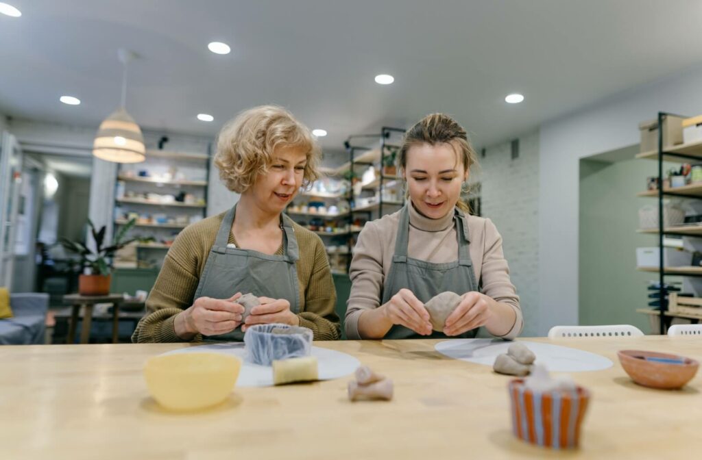 A senior learns from a professional pottery artist on how to model clay in a well-lit art studio