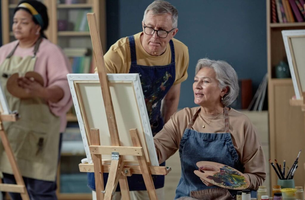 A senior receiving help from an instructor in an art therapy class sitting at an easel with a paintbrush