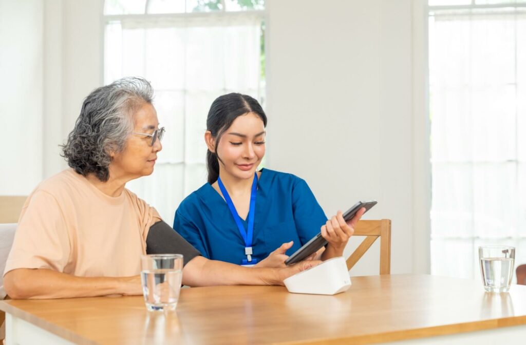 A caregiver sits beside an older adult in a warm kitchen and shows them their medical results on a handheld tablet after a quick checkup