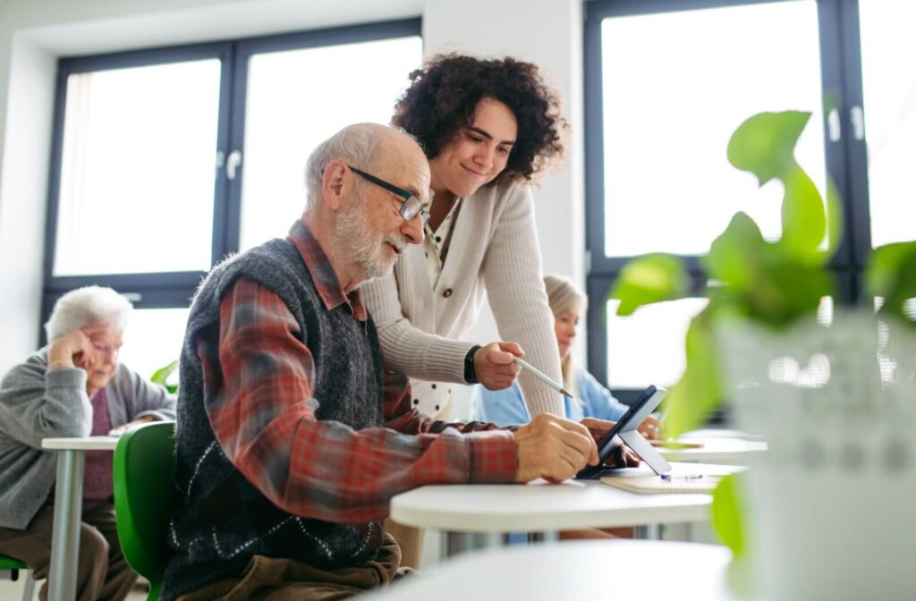 A caregiver in memory care uses a stylus to point at a tablet while helping a smiling resident learn something during a class