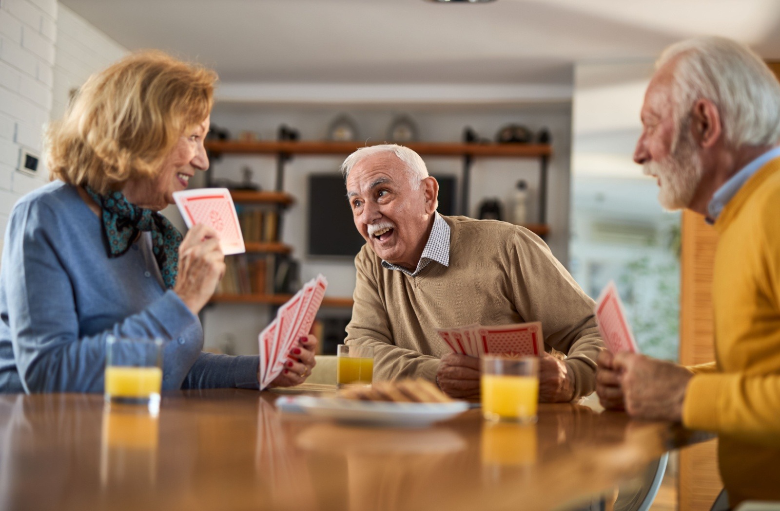 a group of senior friends play a game of cards while enjoying cookies and orange juice.

